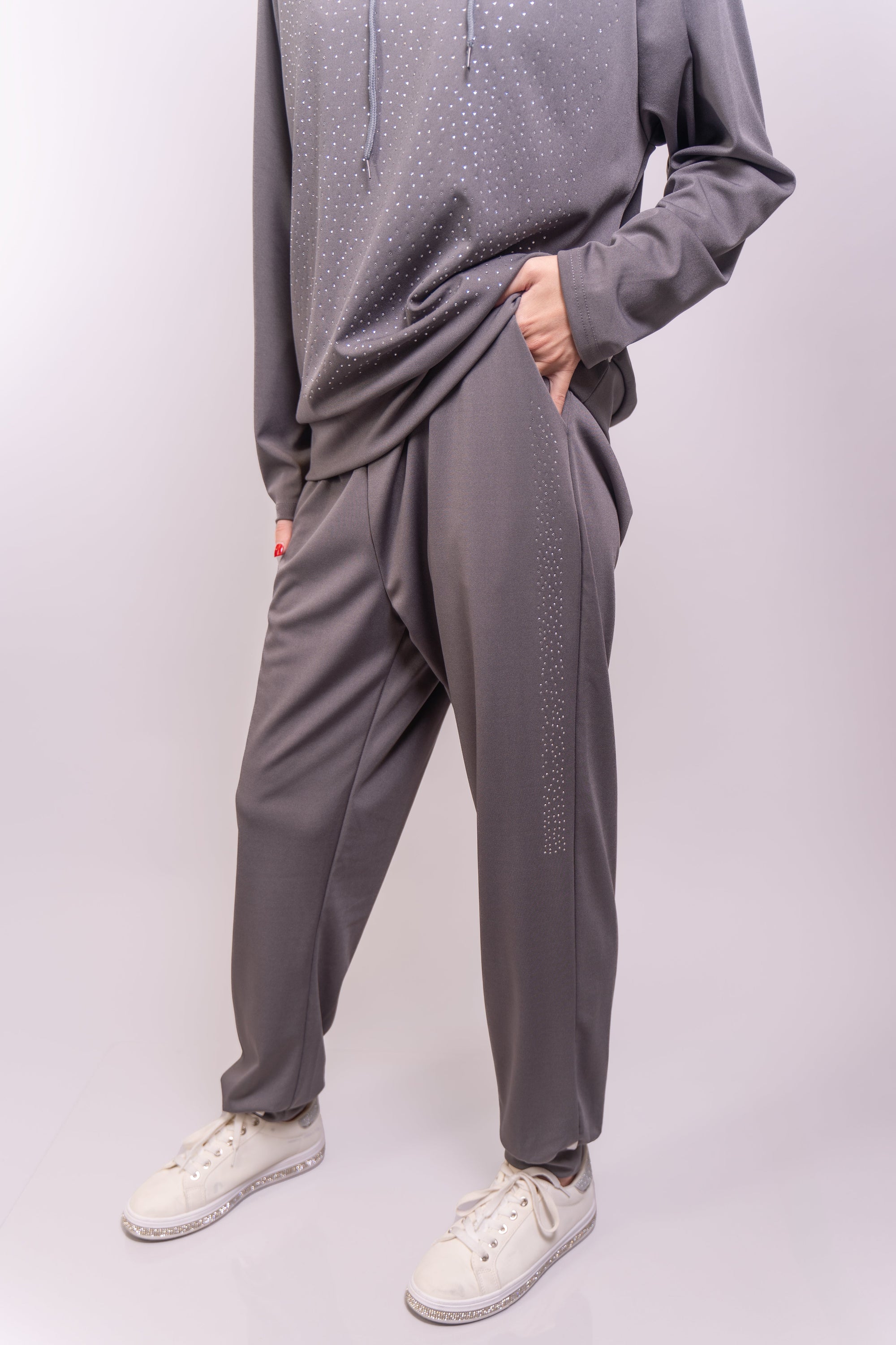 A close up, front-facing view of our model, Lexi, wearing a charcoal gray colored rhinestone embellished hoodie and sweatpants set, focusing on the pants. The outfit is paired with white rhinestone sneakers, also available online and in store.