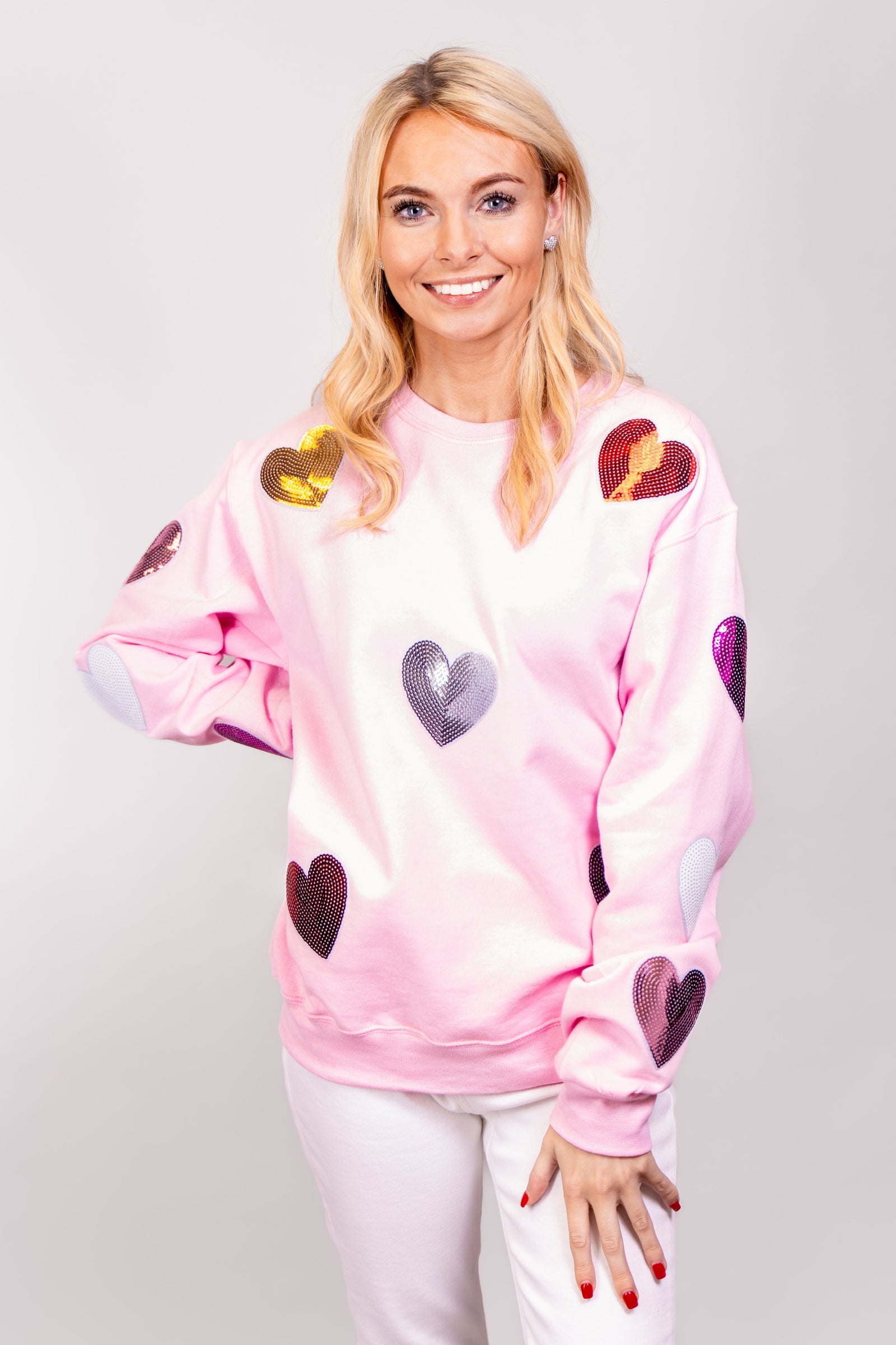 A front view of our model, Lexi, wearing a baby pink sweatshirt covered in Pink, Magenta, Red, Silver and Gold sequin hearts.