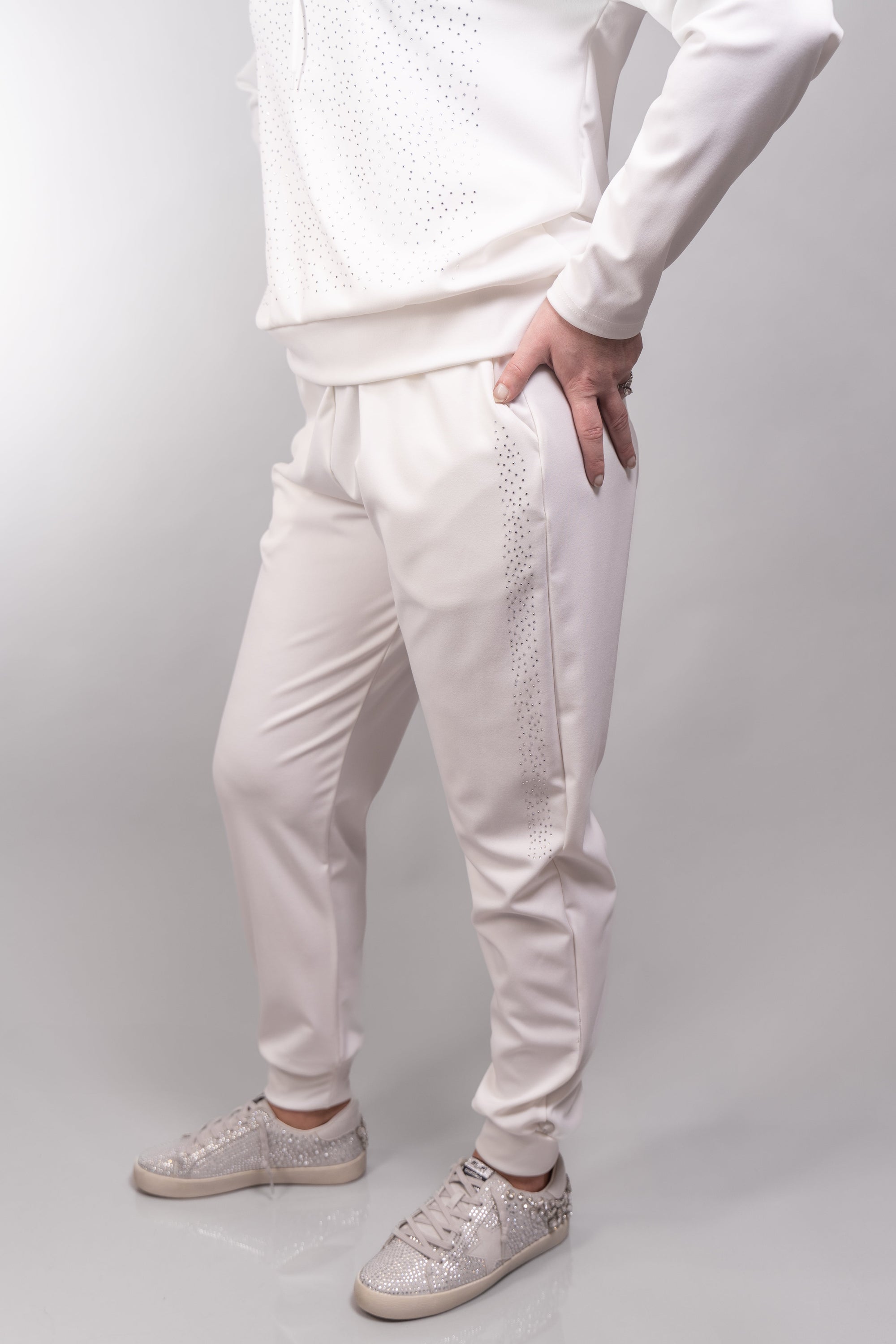 A close-up, side view of our model, Stephanie, wearing a white rhinestone embellished hoodie and sweatpants set, focusing on the pants. This outfit is paired with white rhinestone sneakers, also available online and in store.