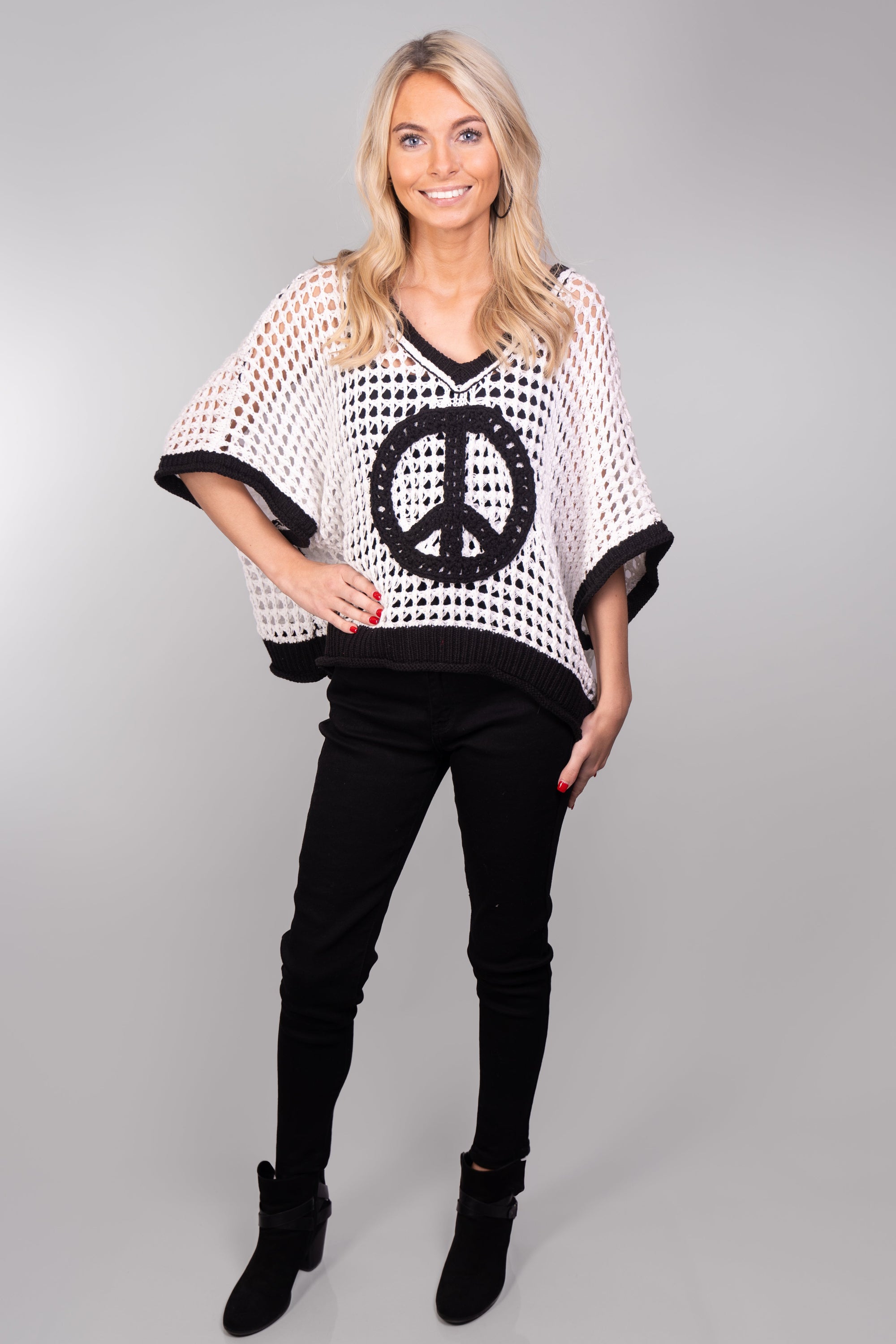 A front-facing, full body view of our model, Lexi, wearing an Ivory loose-knitted short-sleeve oversized top with black trim and a large black peace symbol on the front. This top is pictured and pairs well with black skinny jeans, black heeled ankle boots, and black rhinestone hoops. The black rhinestone hoops are also available for sale online and in store.