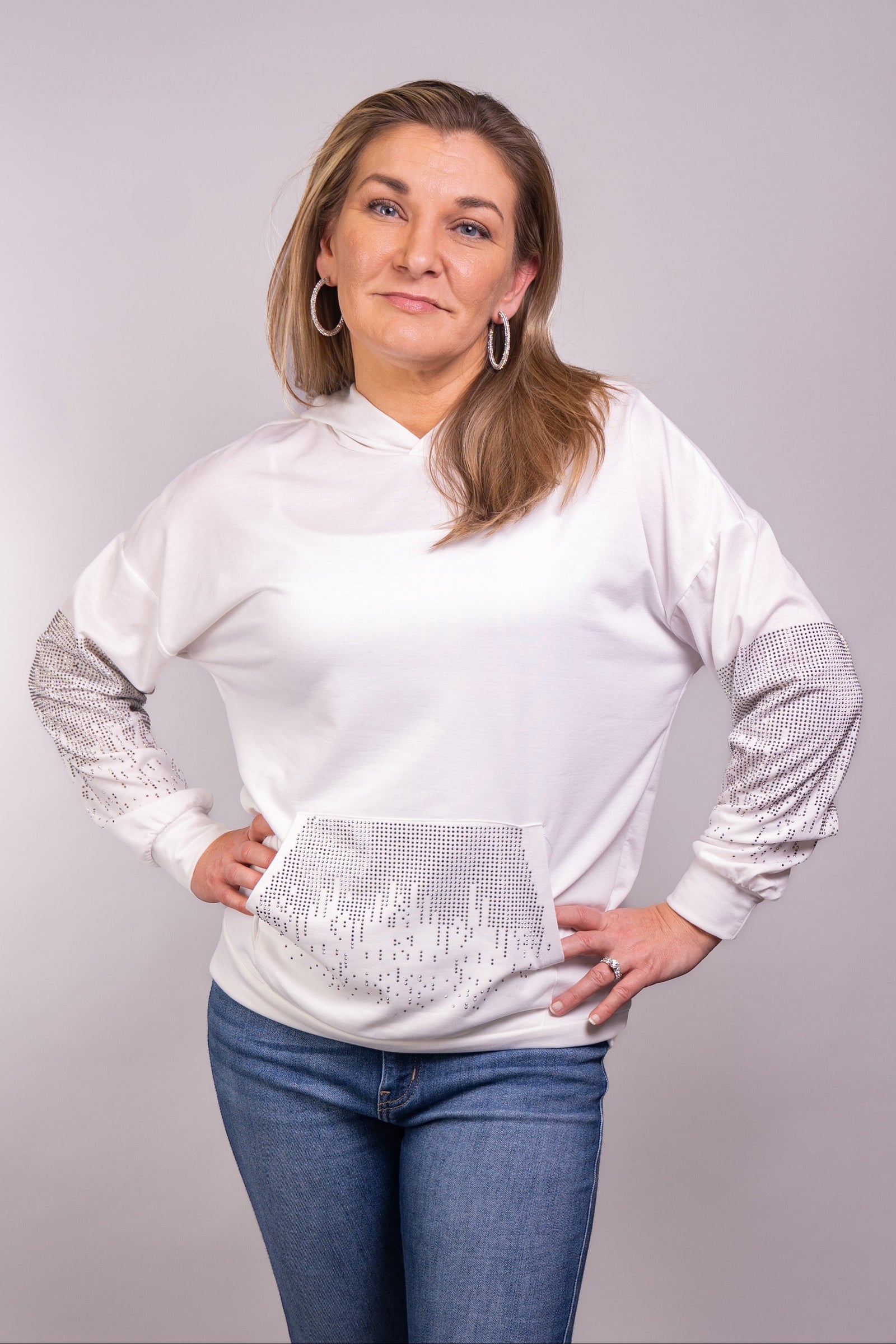 A close-up, front-facing view of our model, Stephanie, wearing a white hoodie embellished with small silver rhinestones in a dripping pattern on the sleeves and front pocket. Pictured here paired nicely with light wash skinny jeans.