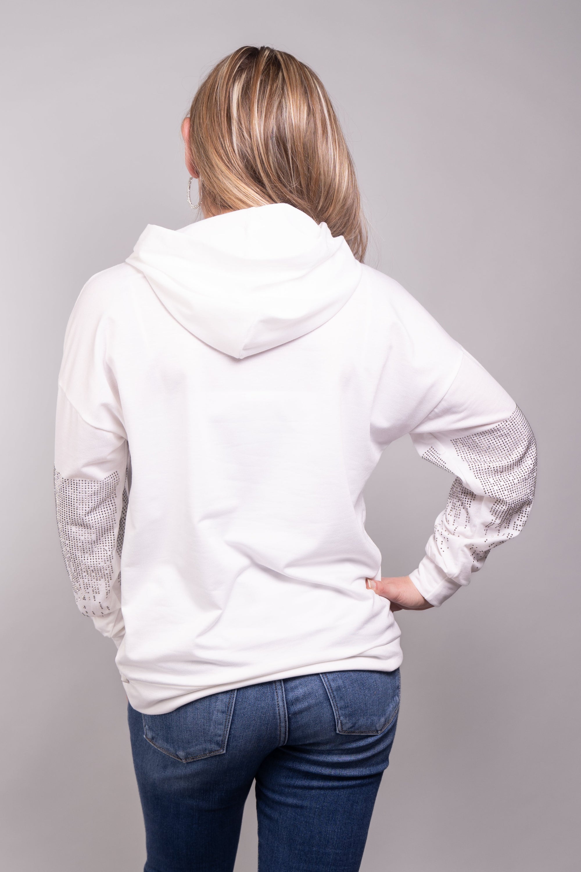 A close-up, front-facing view of our model, Stephanie, wearing a white hoodie embellished with small silver rhinestones in a dripping pattern on the sleeves and front pocket. Pictured here paired nicely with light wash skinny jeans.