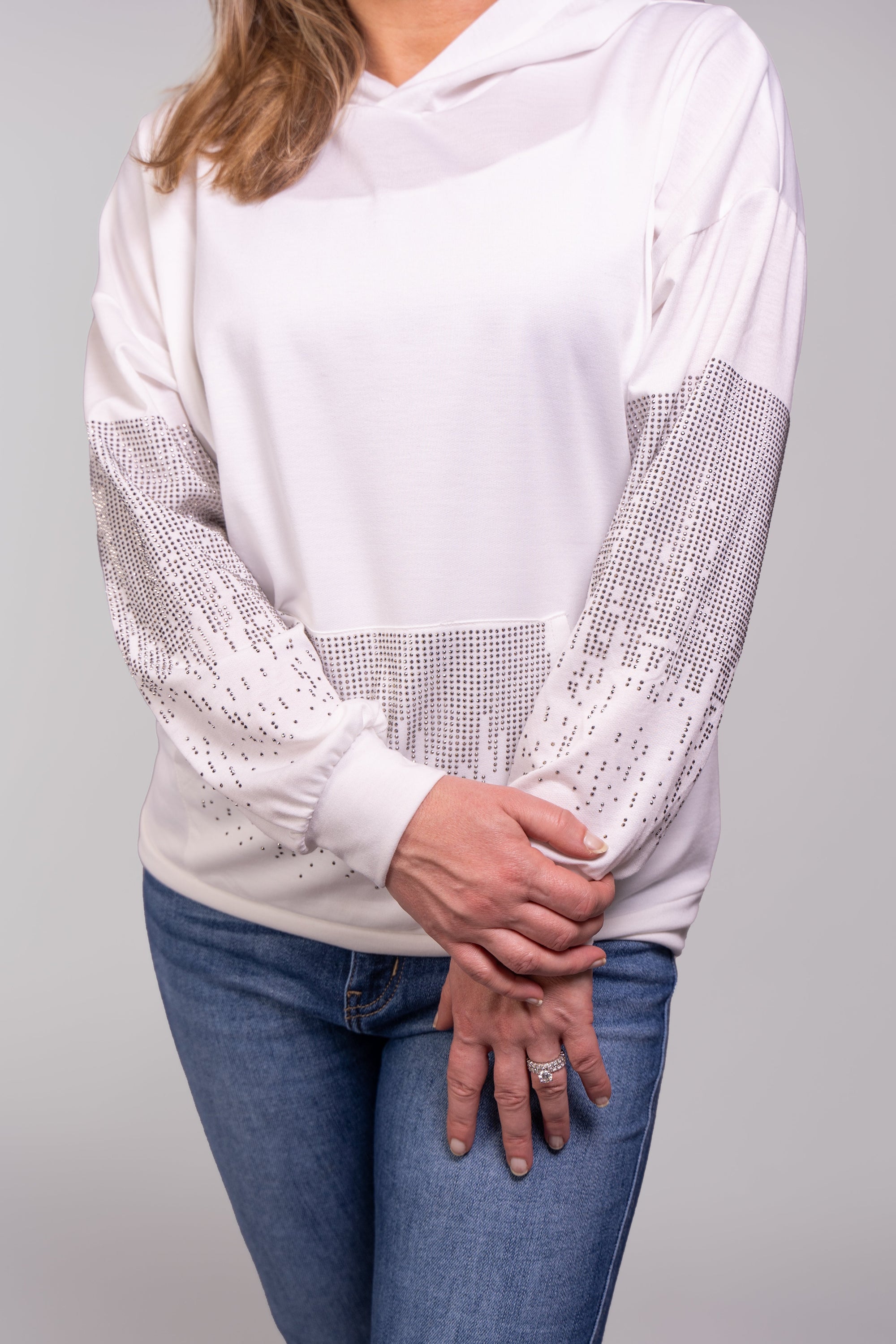 A close-up, front-facing view of our model, Stephanie, wearing a white hoodie embellished with small silver rhinestones in a dripping pattern on the sleeves and front pocket. Pictured here paired nicely with light wash skinny jeans. This photo is zoomed in on the sleeve and pocket details, showing off the rhinestones.