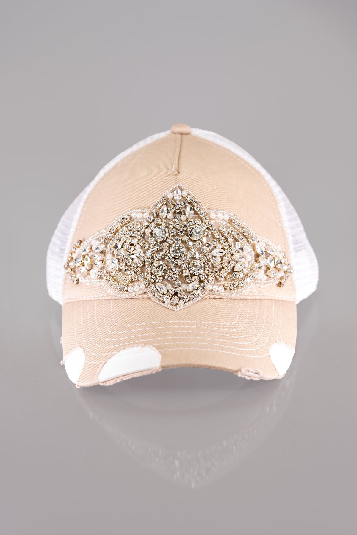 A front facing view of a beige and white trucker style baseball cap with distressed bill and a bold embellishment of rhinestones and pearls on the front.