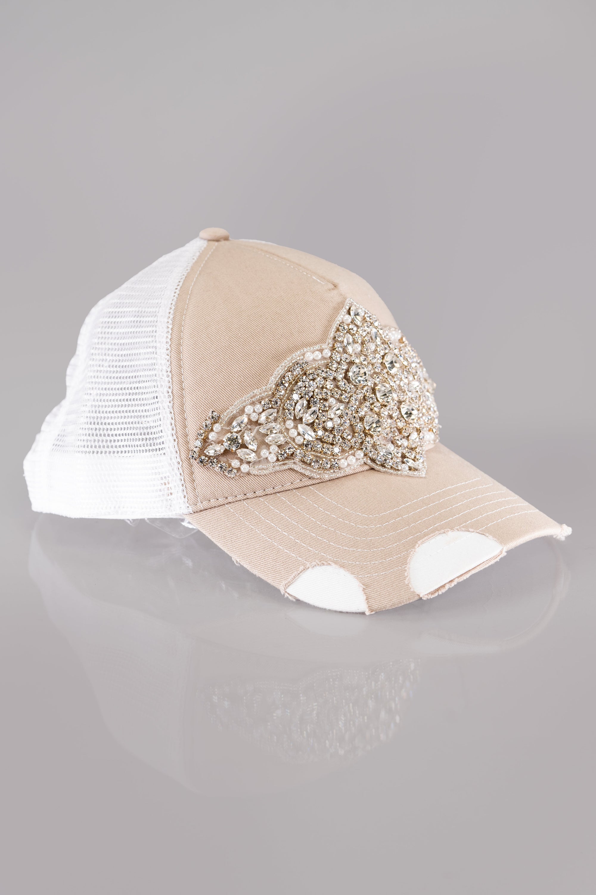 A side view of a beige and white trucker style baseball cap with distressed bill and a bold embellishment of rhinestones and pearls on the front.