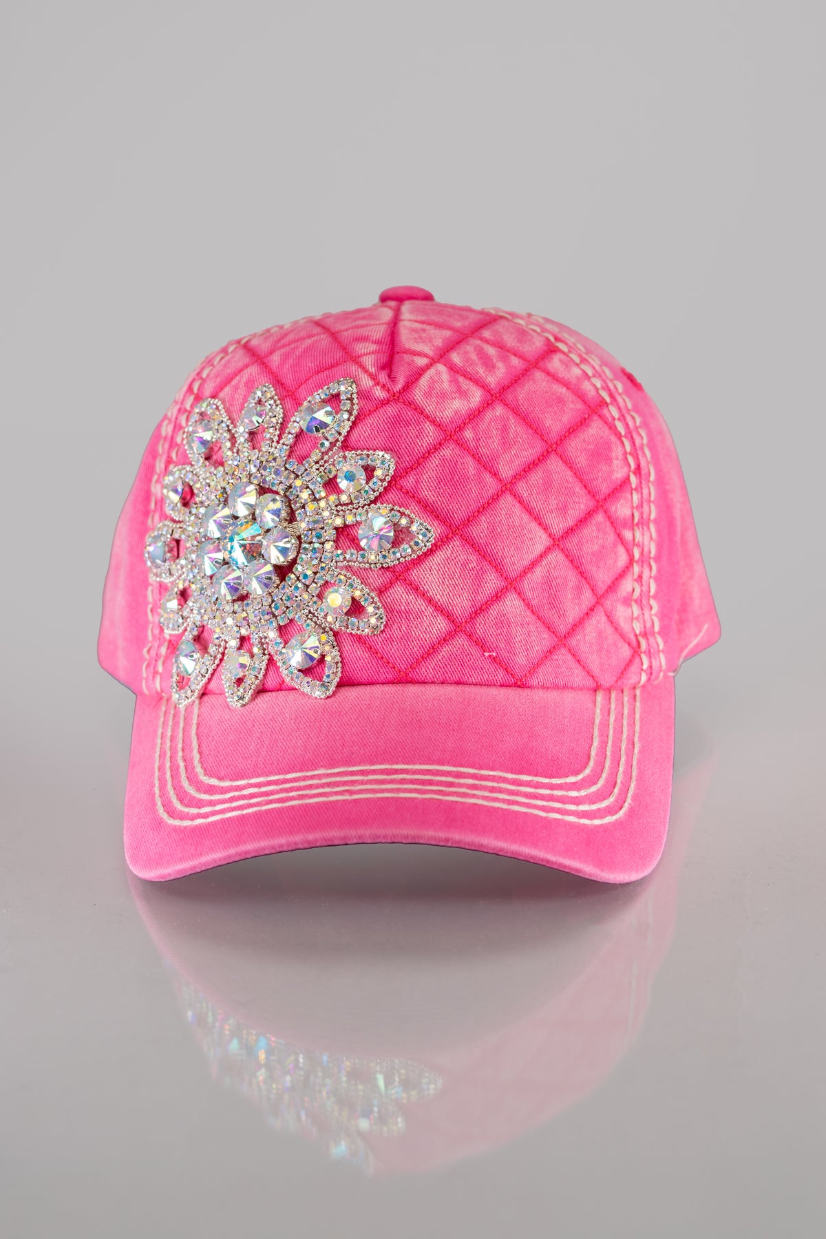 A front facing view of a bright pink quilted baseball cap with a large rhinestone flower embellished on the front and white stitching detail throughout.