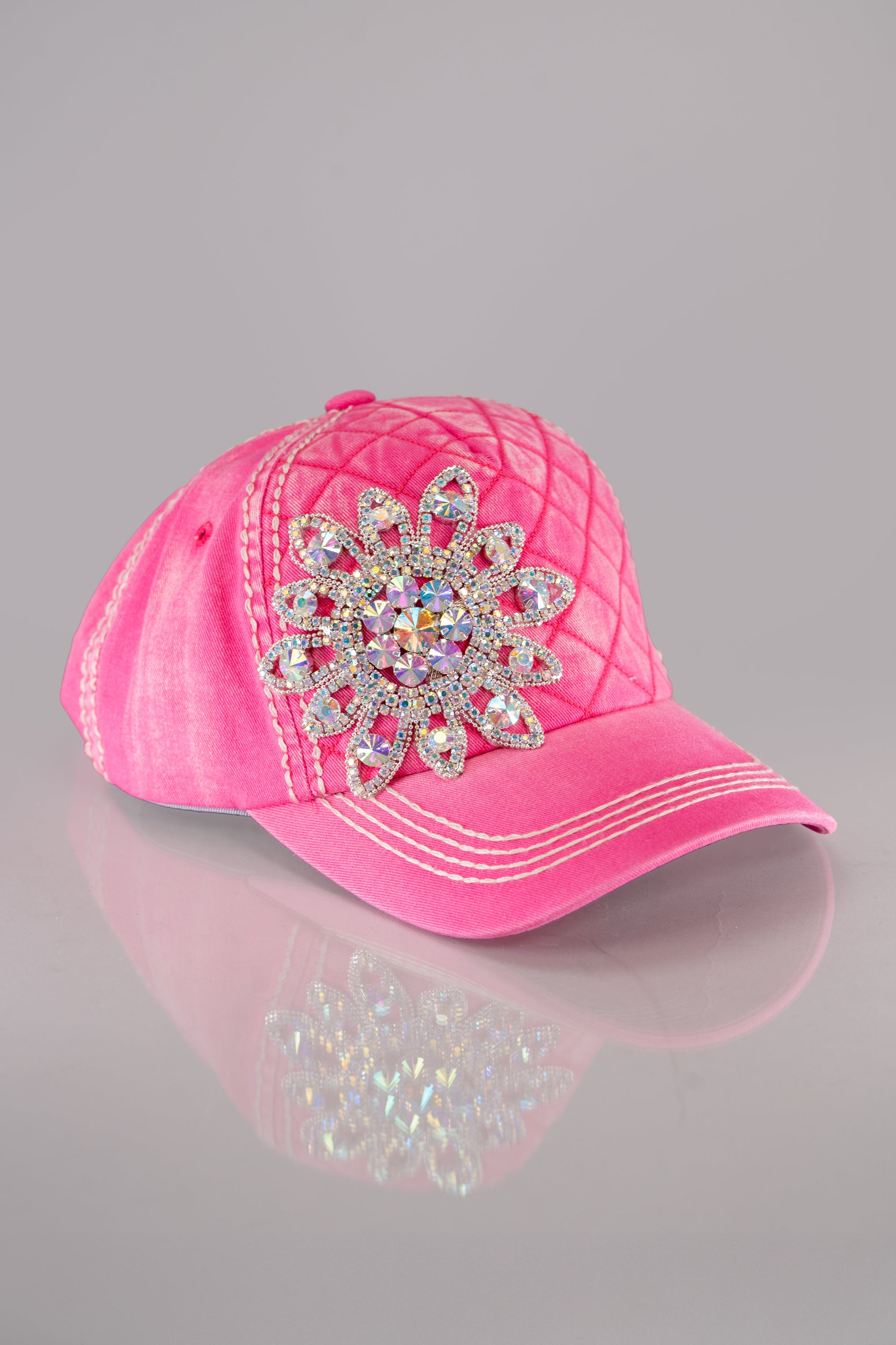 A side view of a bright pink quilted baseball cap with a large rhinestone flower embellished on the front and white stitching detail throughout.