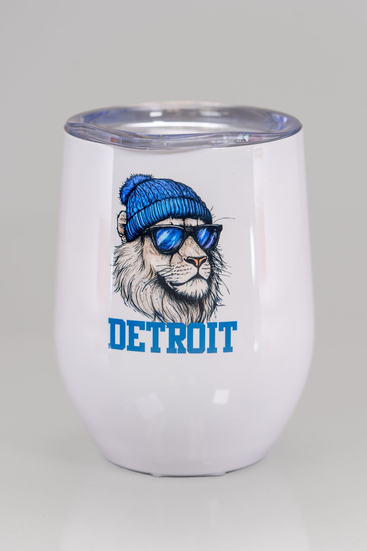 A white stainless steel tumbler with a design on the front featuring a lion wearing a blue pom beanie and black sunglasses. Beneath the lion it says &#39;DETROIT&#39; in a bold blue serif font.