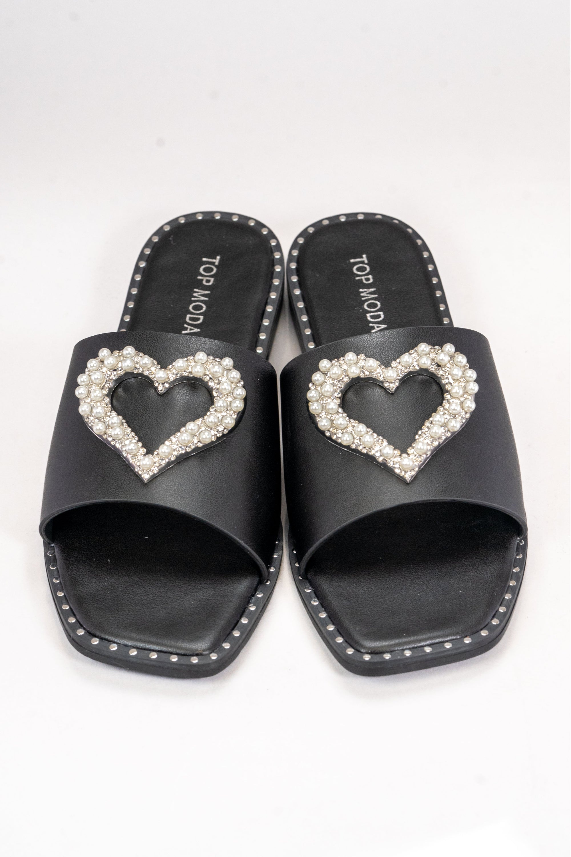 A front-facing view of a pair of black slide sandals with silver studs lining the perimeter of the shoes. This style also features a large heart on top made up of rhinestone and pearl embellishment. In this picture one shoe is lightly resting on the other.