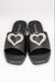 A front-facing view of a pair of black slide sandals with silver studs lining the perimeter of the shoes. This style also features a large heart on top made up of rhinestone and pearl embellishment. In this picture one shoe is lightly resting on the other.