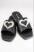 A front-facing view of a pair of black slide sandals with silver studs lining the perimeter of the shoes. This style also features a large heart on top made up of rhinestone and pearl embellishment. In this picture one shoe is lightly resting on the other.