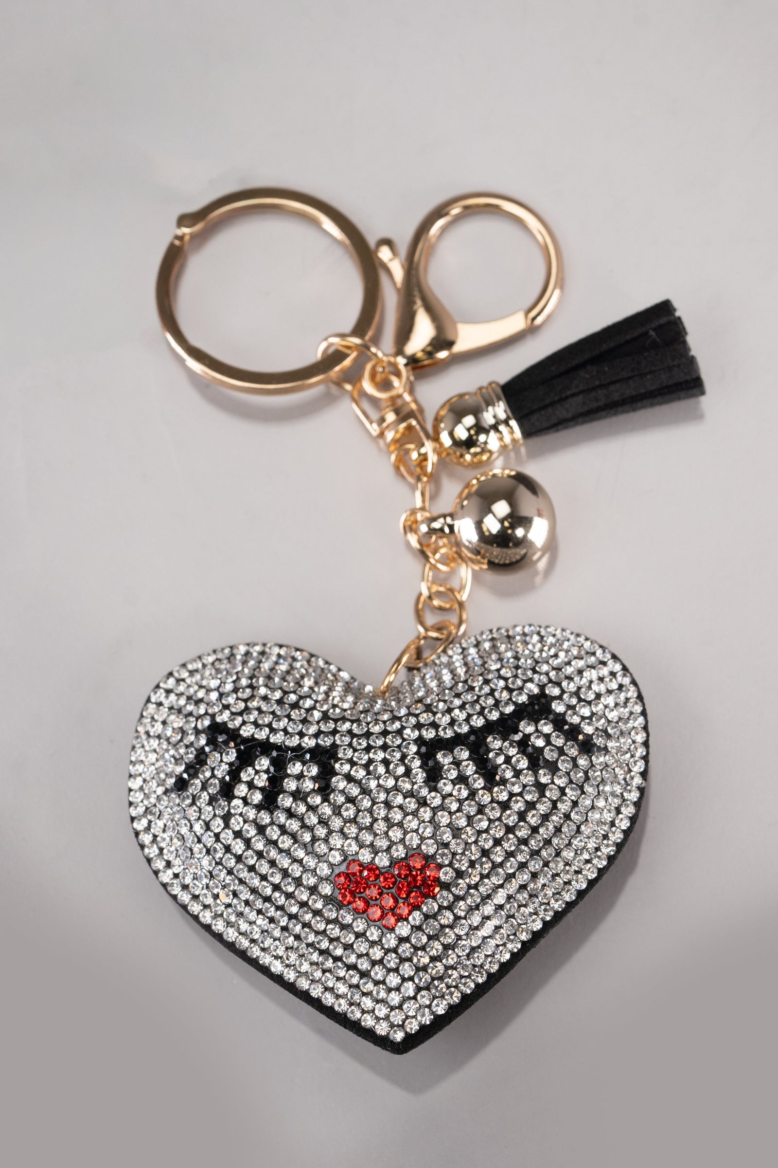 A front view of a black puffy heart keychain with silver rhinestone embellishment and a cute face design on the front, made up of black rhinestone eyelashes and red rhinestone lips in the shape of a heart. This keychain also has gold hardware.