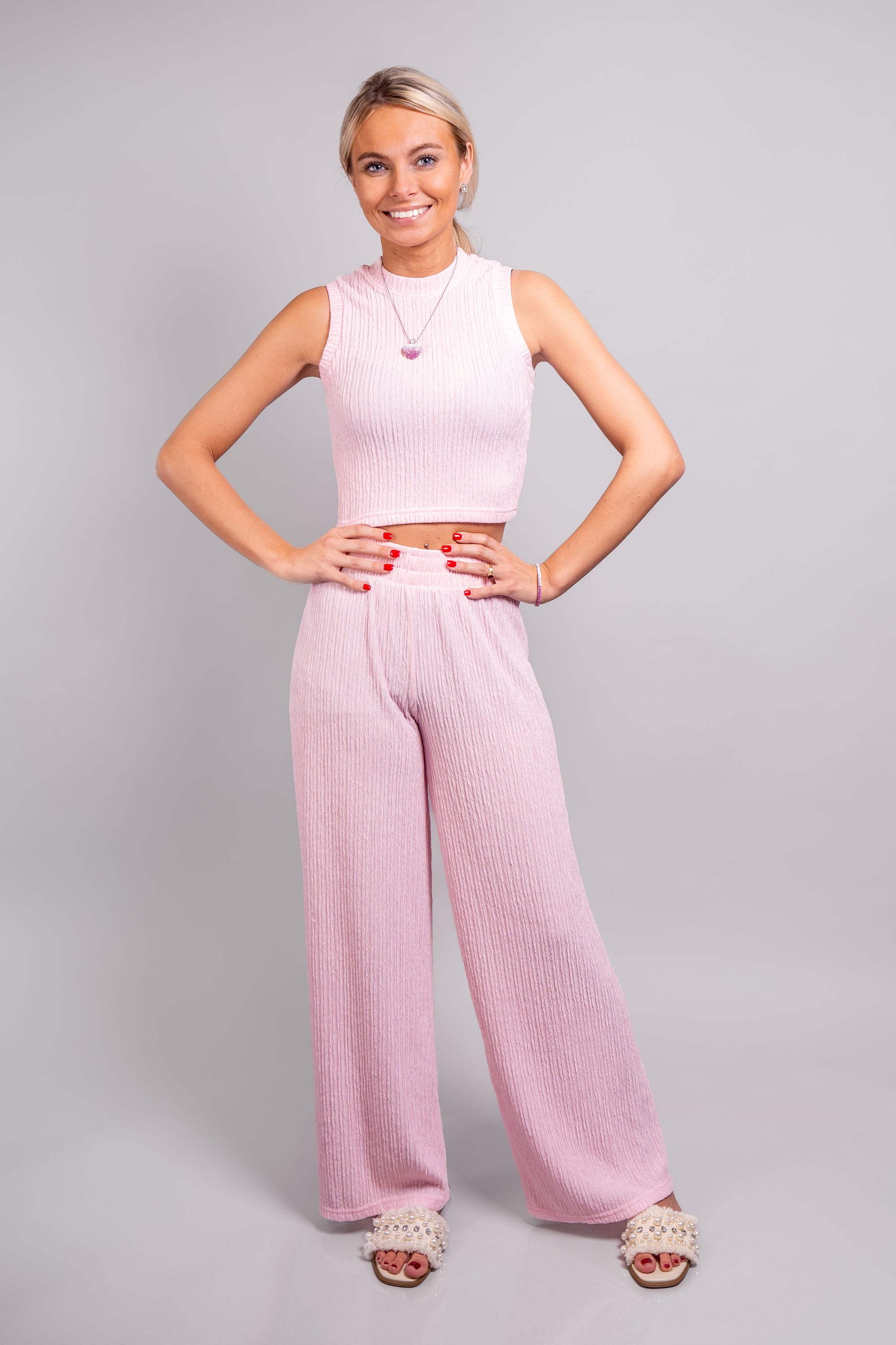 A full body, side view of our model, Lexi, wearing the crinkled sleeveless cropped top and wide leg pants set in light pink. This set is paired with a pink and white ombre rhinestone heart necklace, silver rhinestone earrings, and ivory slide sandals with pearl and rhinestone embellishments.