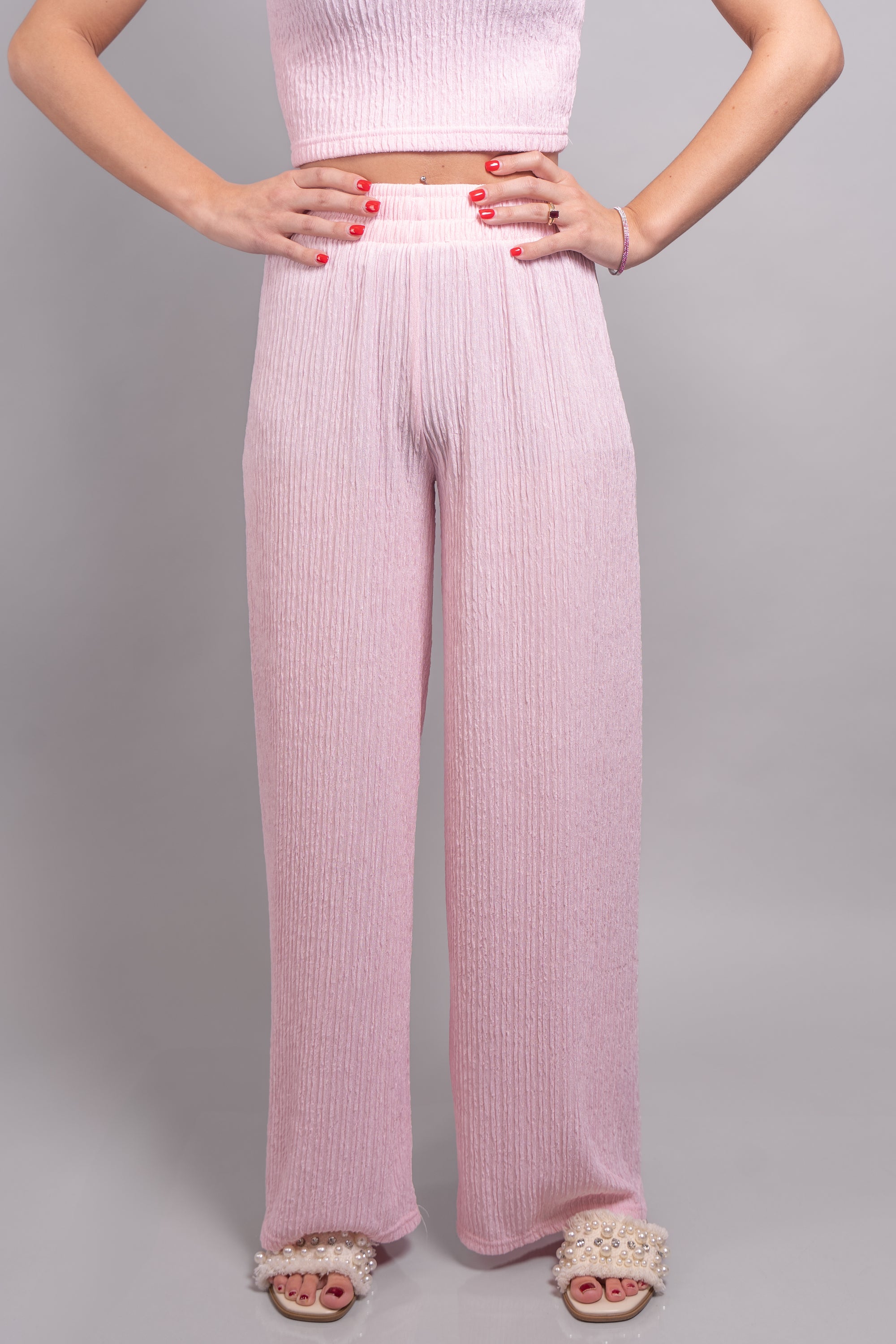 A close-up, front-facing view of our model, Lexi, wearing the crinkled sleeveless cropped top and wide leg pants set in light pink. This set is paired with ivory slide sandals with pearl and rhinestone embellishments. This view is focused on the pants, showing the length of the pant legs, and the detail of the crinkle fabric.