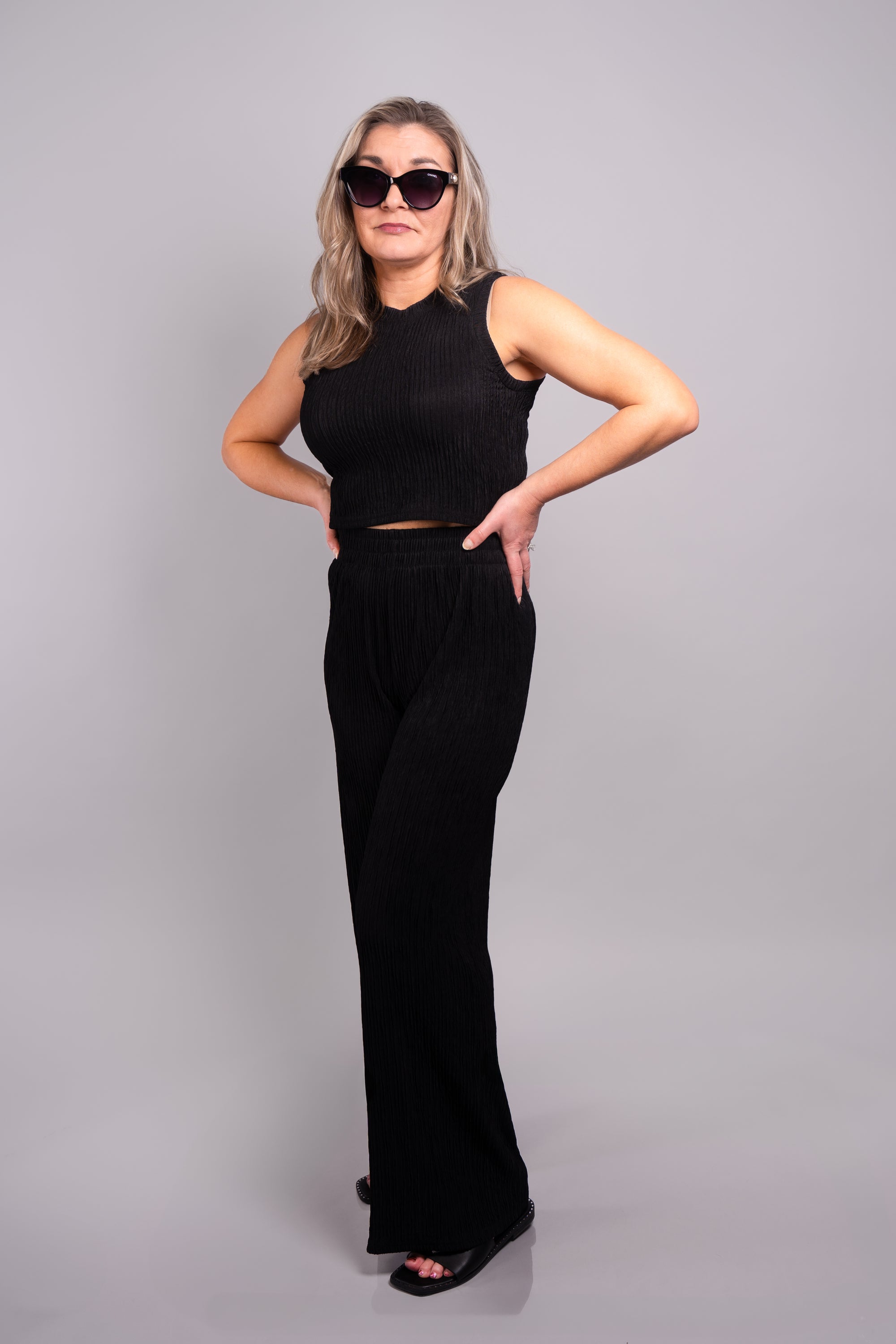 A full body side view of our model, Stephanie, wearing the crinkled sleeveless cropped top and wide leg pants set in black. This set is paired with pearl earrings, cat eye sunglasses, and black slide sandals. 