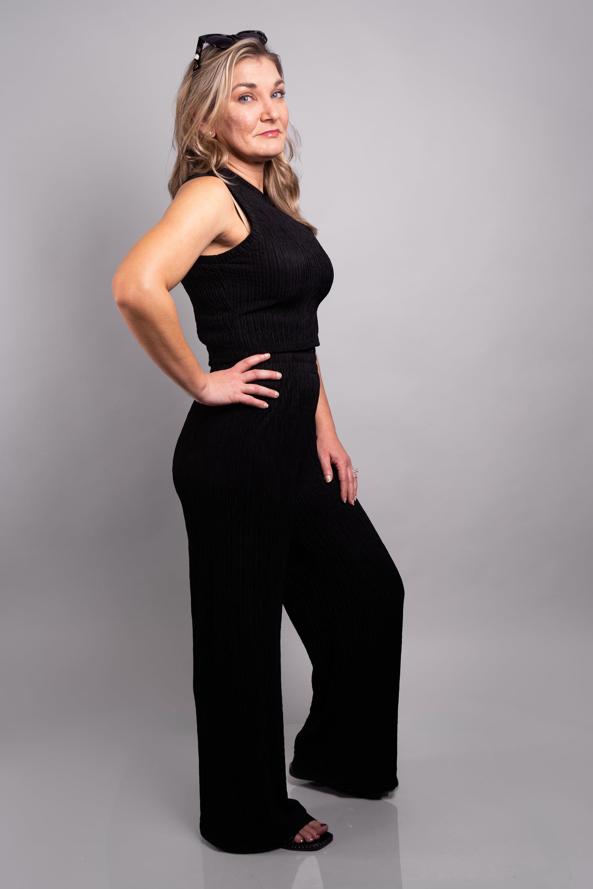 A full body side view of our model, Stephanie, wearing the crinkled sleeveless cropped top and wide leg pants set in black. This set is paired with pearl earrings, cat eye sunglasses, and black slide sandals.