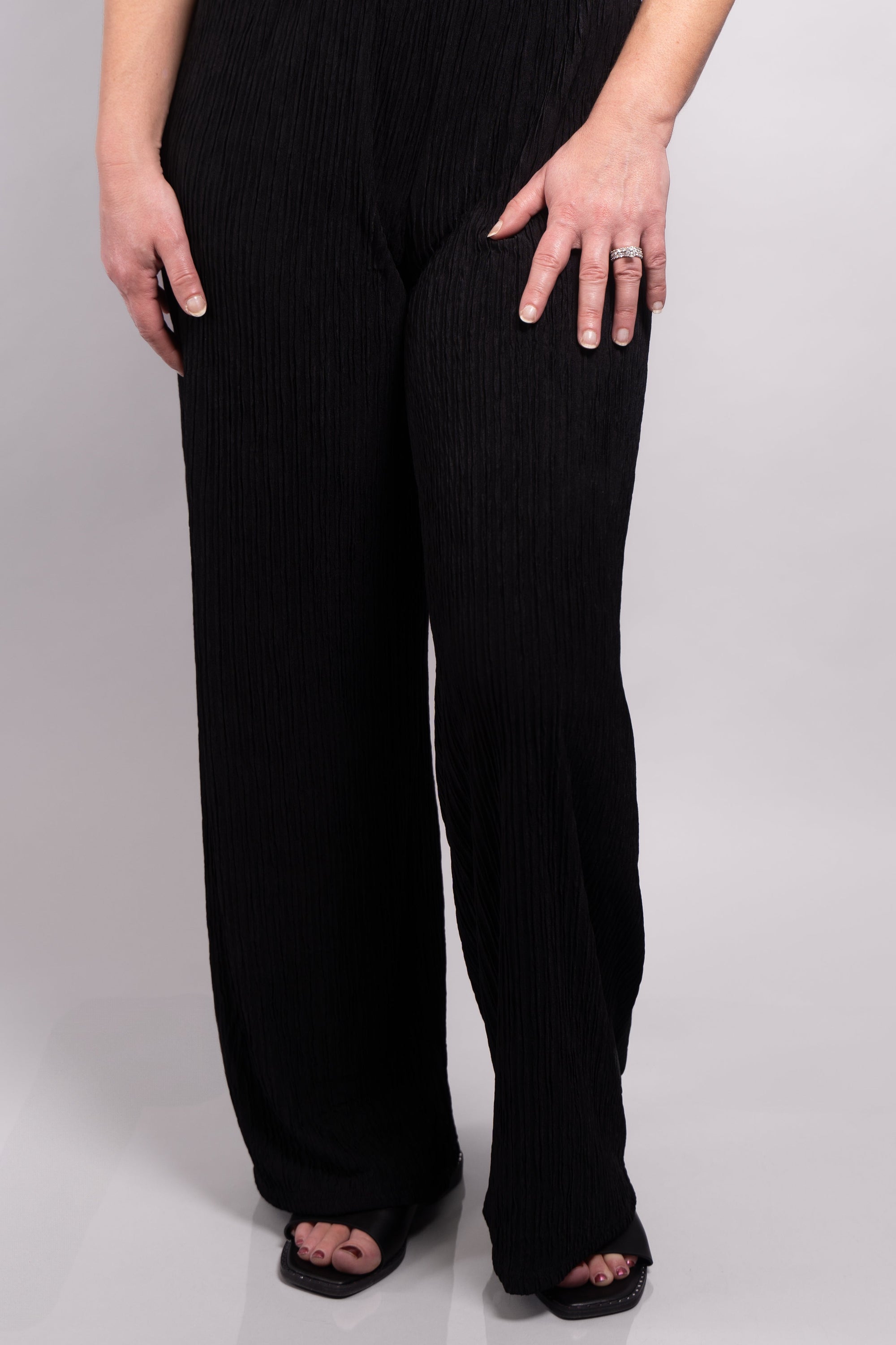 A close-up front-facing view of our model, Stephanie, wearing the crinkled sleeveless cropped top and wide leg pants set in black. This set is paired with black slide sandals. This photo is focused on the pants, showing the length of the pant legs and the detail of the unique crinkle fabric.