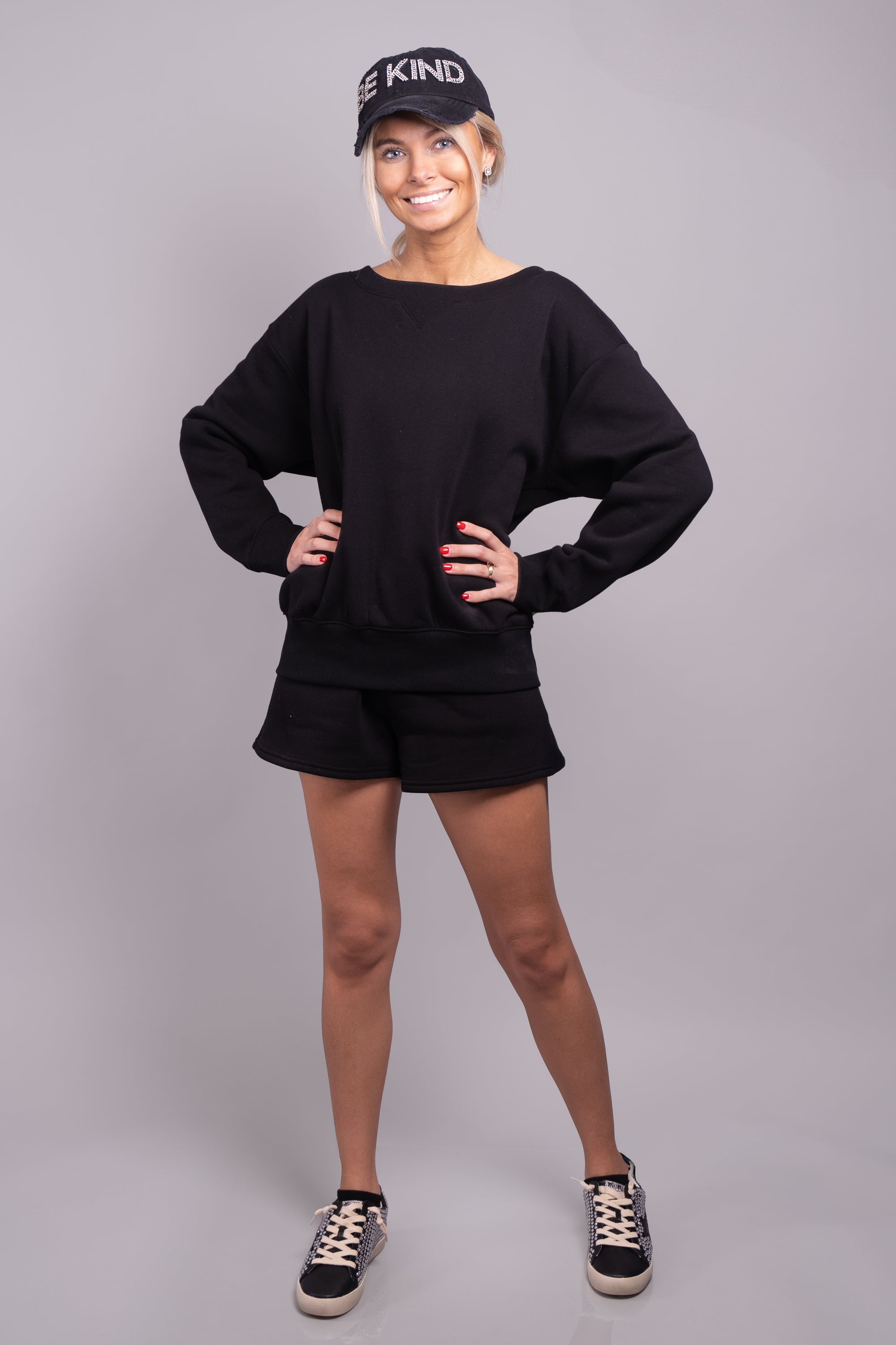 A front-facing full body view of our model, Lexi, wearing a fleece sweatshirt and shorts set, in Black. The set is paired with a black rhinestone baseball hat and black and ivory rhinestone sneakers.