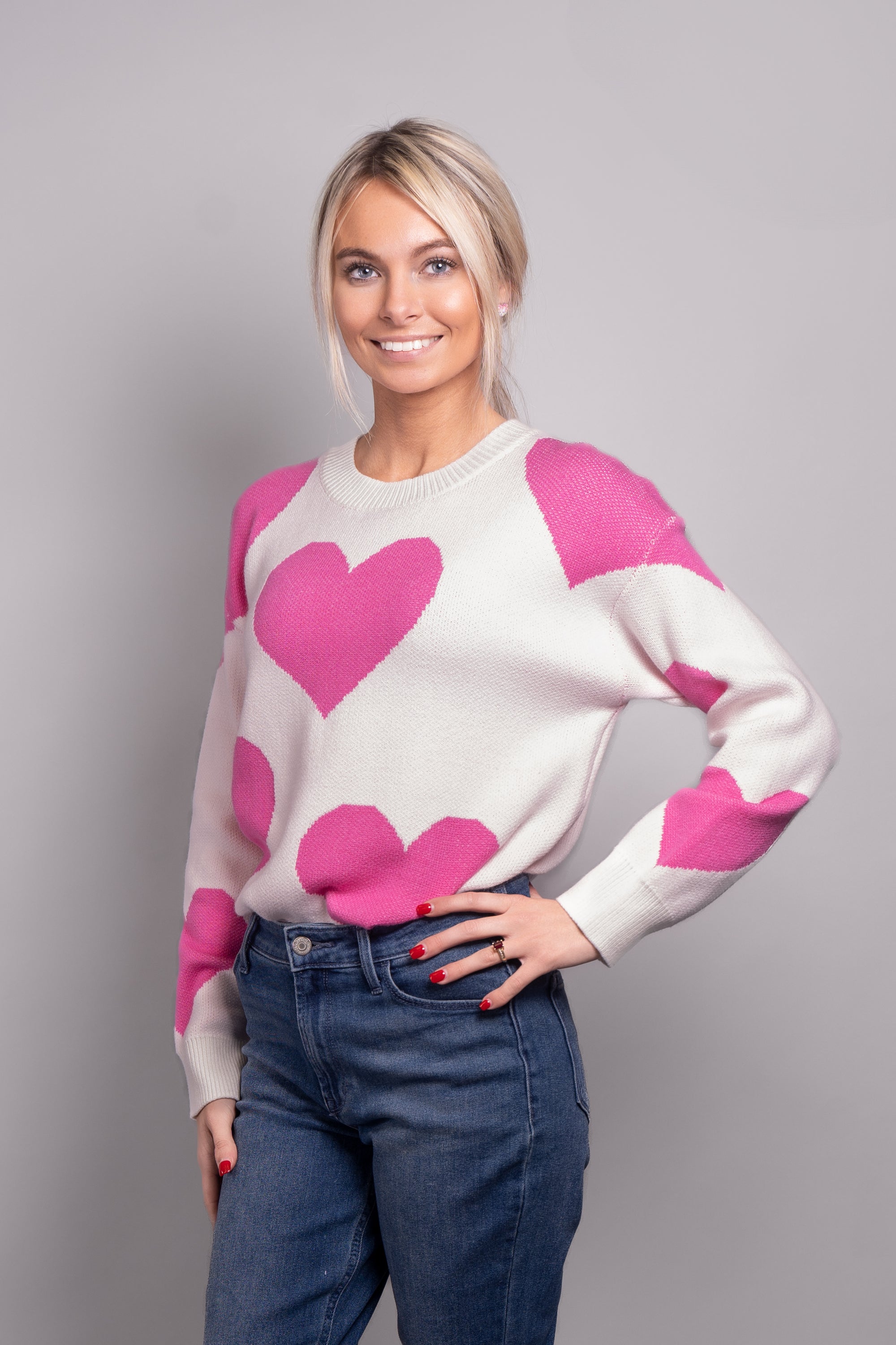 A close-up, side view of our model, Lexi, wearing a white crew neck sweater covered in large pink hearts all over. Paired with blue jeans, and pink and white ombre rhinestone heart earrings for a casual look.