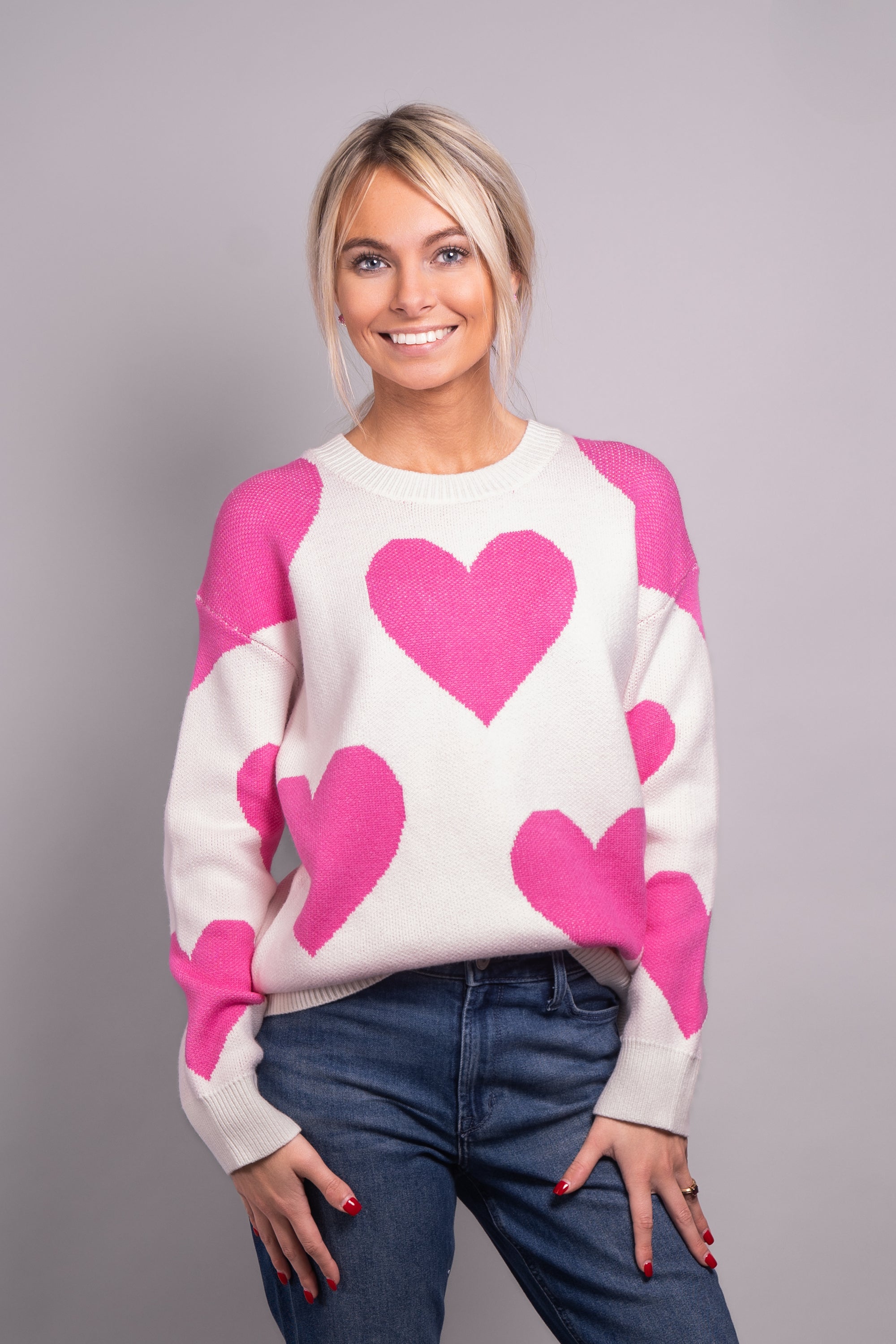 A close-up, front-facing view of our model, Lexi, wearing a white crew neck sweater covered in large pink hearts all over. Paired with blue jeans,  and pink and white ombre rhinestone heart earrings for a casual look.