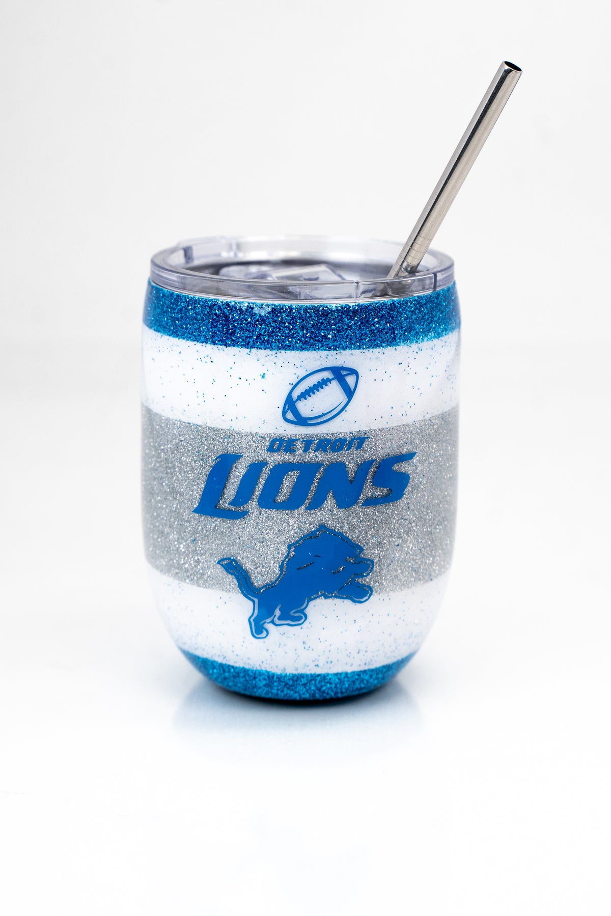 A stainless steel tumblr with stainless steel straw and a clear top. The design features alternating stripes of white, blue and silver glitter, with the Detroit Lions logos on the front.